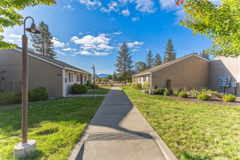 A concrete pathway runs between two single-story beige buildings with green lawns and trees under a bright blue sky with scattered clouds. Mountains are visible in the background.