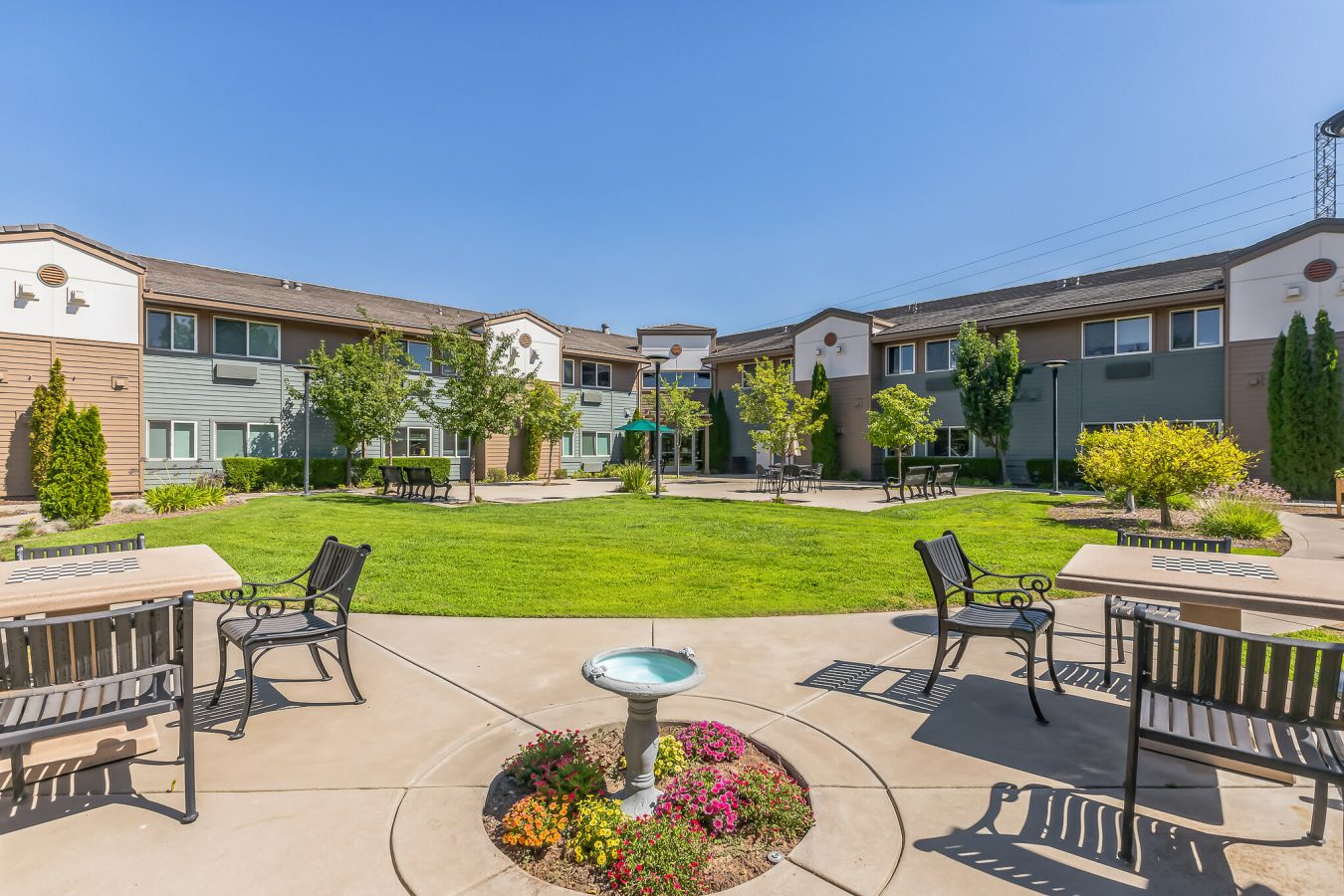 A courtyard with green grass, flowers, and trees, surrounded by two-story apartment buildings. There are benches and tables on the paved area, and a small decorative fountain in the center under a clear blue sky.