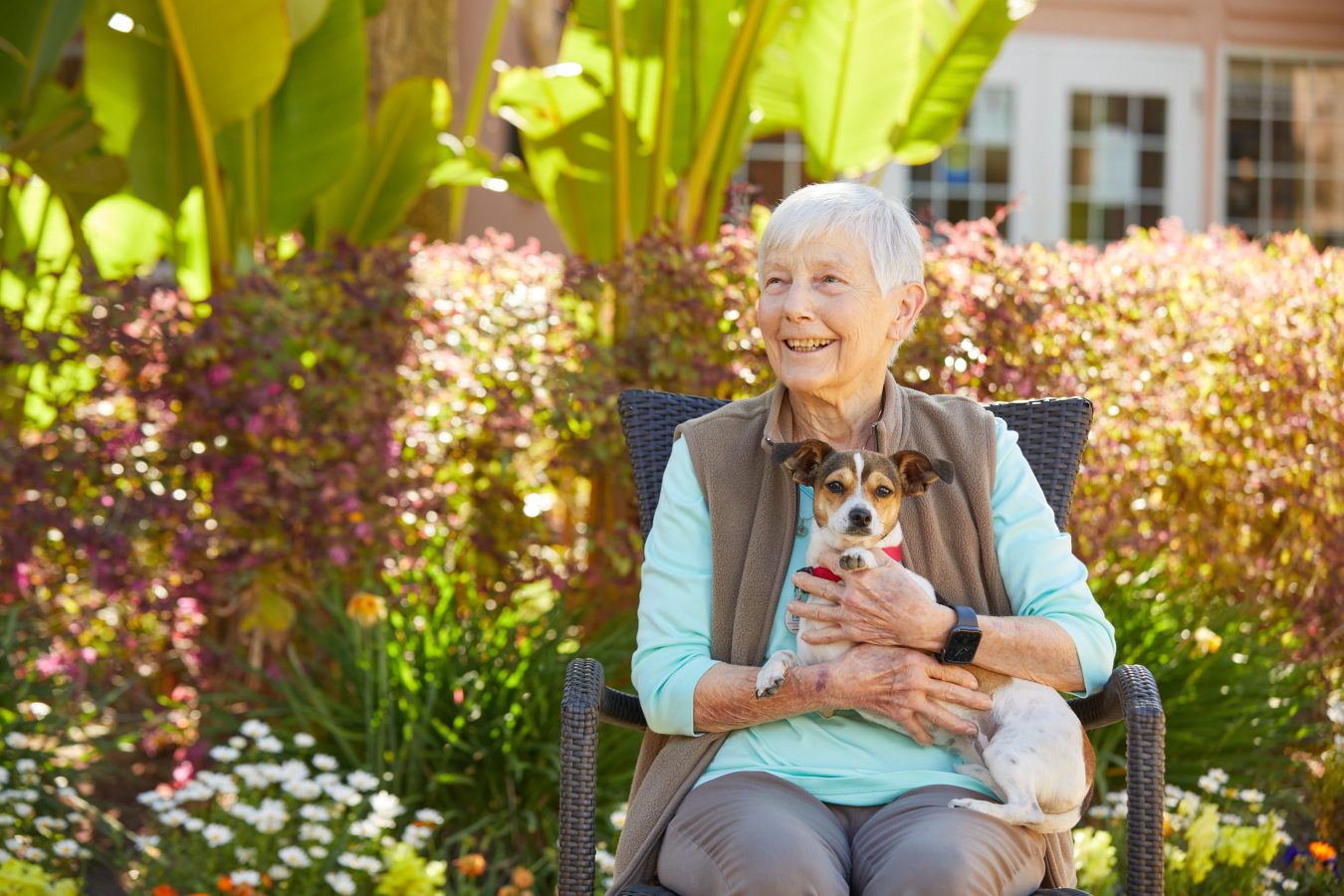 An elderly woman with short gray hair sits outdoors in a garden, smiling and holding a small dog in her lap. She wears a light blue shirt and beige vest, surrounded by lush greenery and flowers.