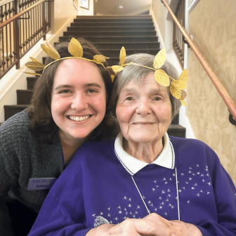 Two women, one younger and one older, smile together indoors. Both wear gold leaf crowns. The younger woman has short brown hair and a nametag, while the older woman wears a purple sweater and sits in a wheelchair.