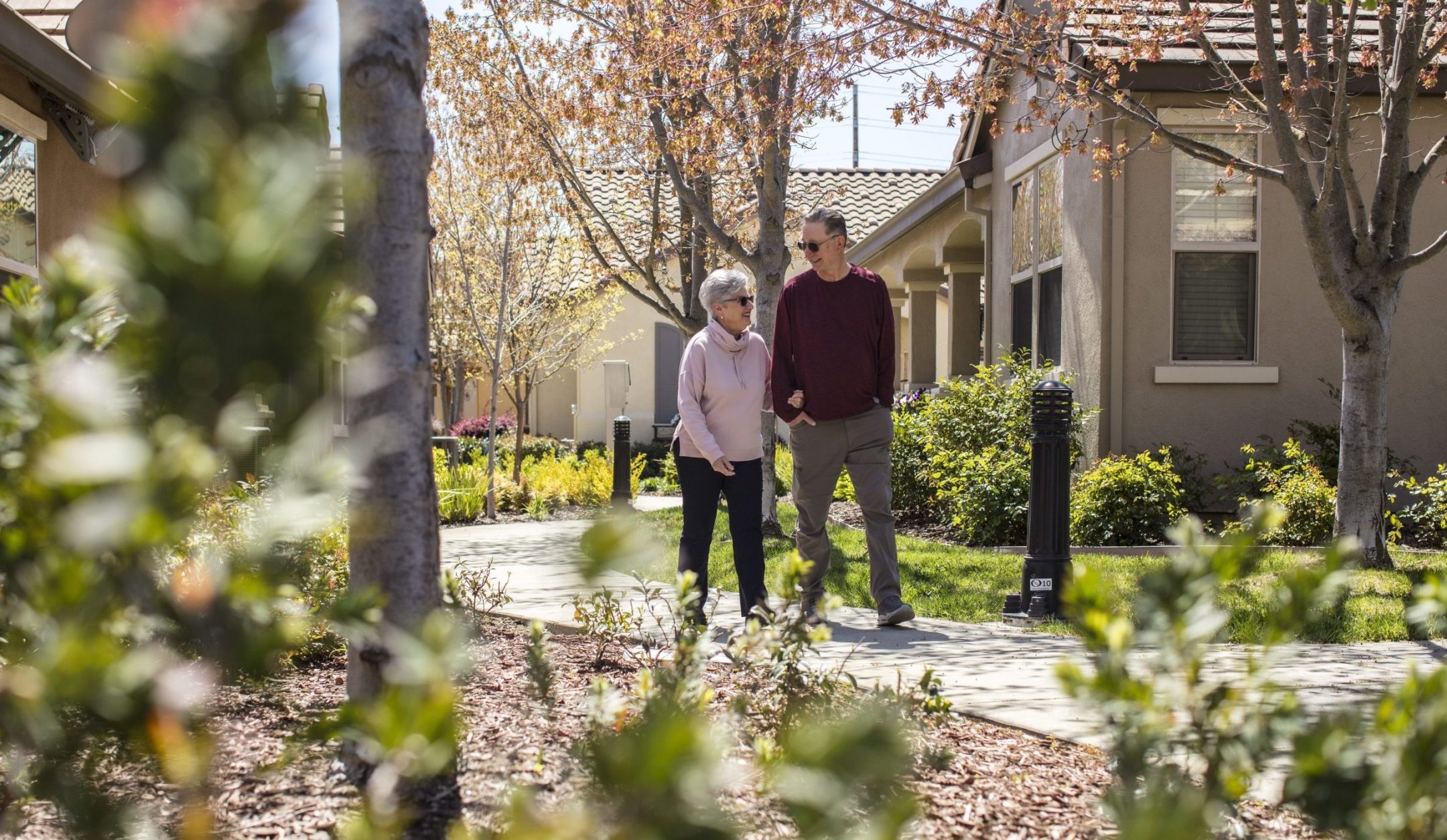 An older couple walks arm in arm along a garden path in a residential neighborhood with trees and greenery, enjoying a sunny day.