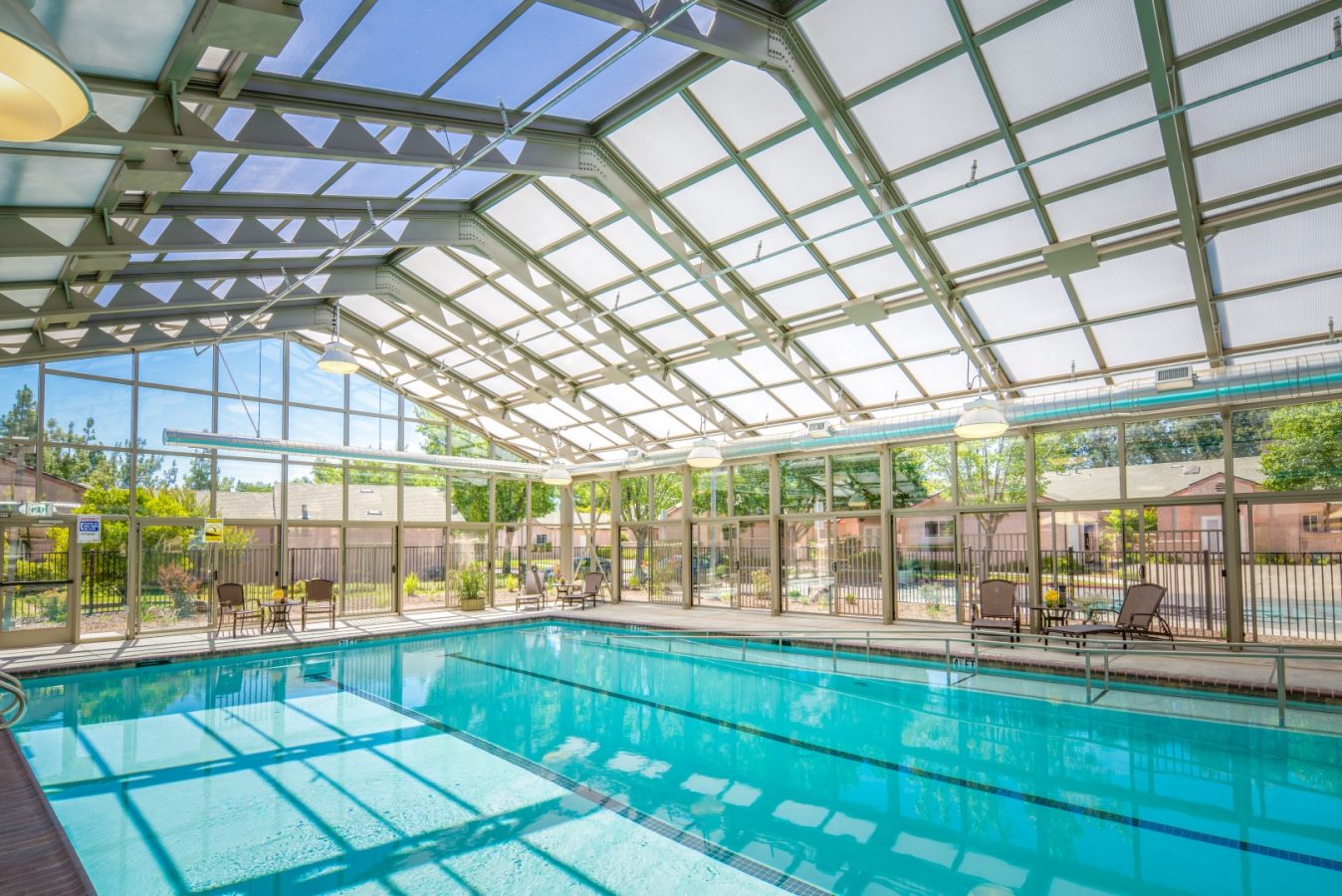 Bright indoor swimming pool with clear blue water, surrounded by lounge chairs and tables; large glass windows and a vaulted glass ceiling let in natural sunlight and offer views of trees and residential buildings outside.