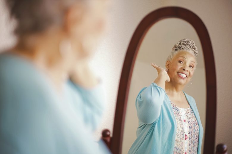 A smiling older woman with gray hair looks at herself in a mirror, wearing a light blue cardigan and patterned blouse, touching her neck with one hand in a warm, softly lit room.