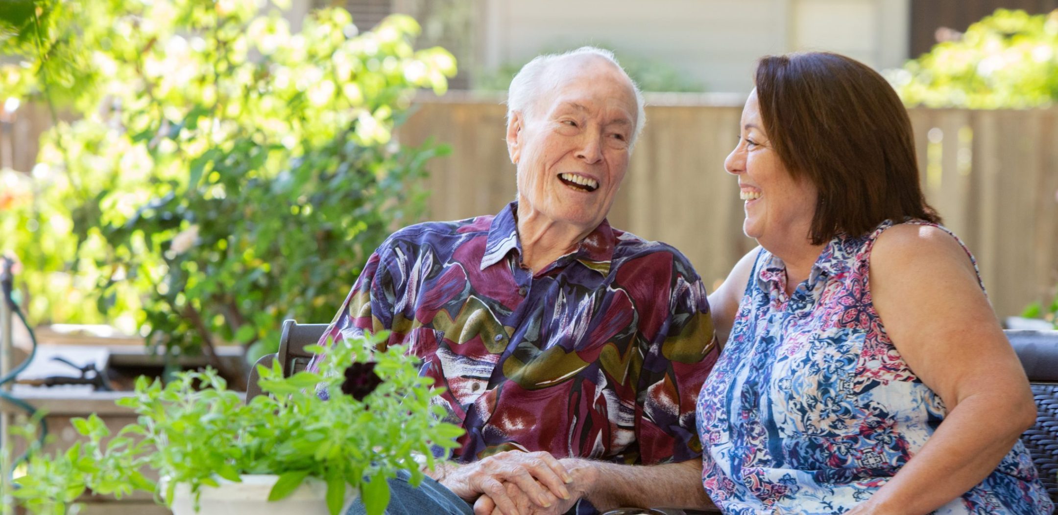 An older man and woman sit outside on patio chairs, smiling and laughing together. A table with a teapot, cups, and potted plants sits in front of them as they enjoy the afternoon, perhaps after attending Eskaton support groups together.