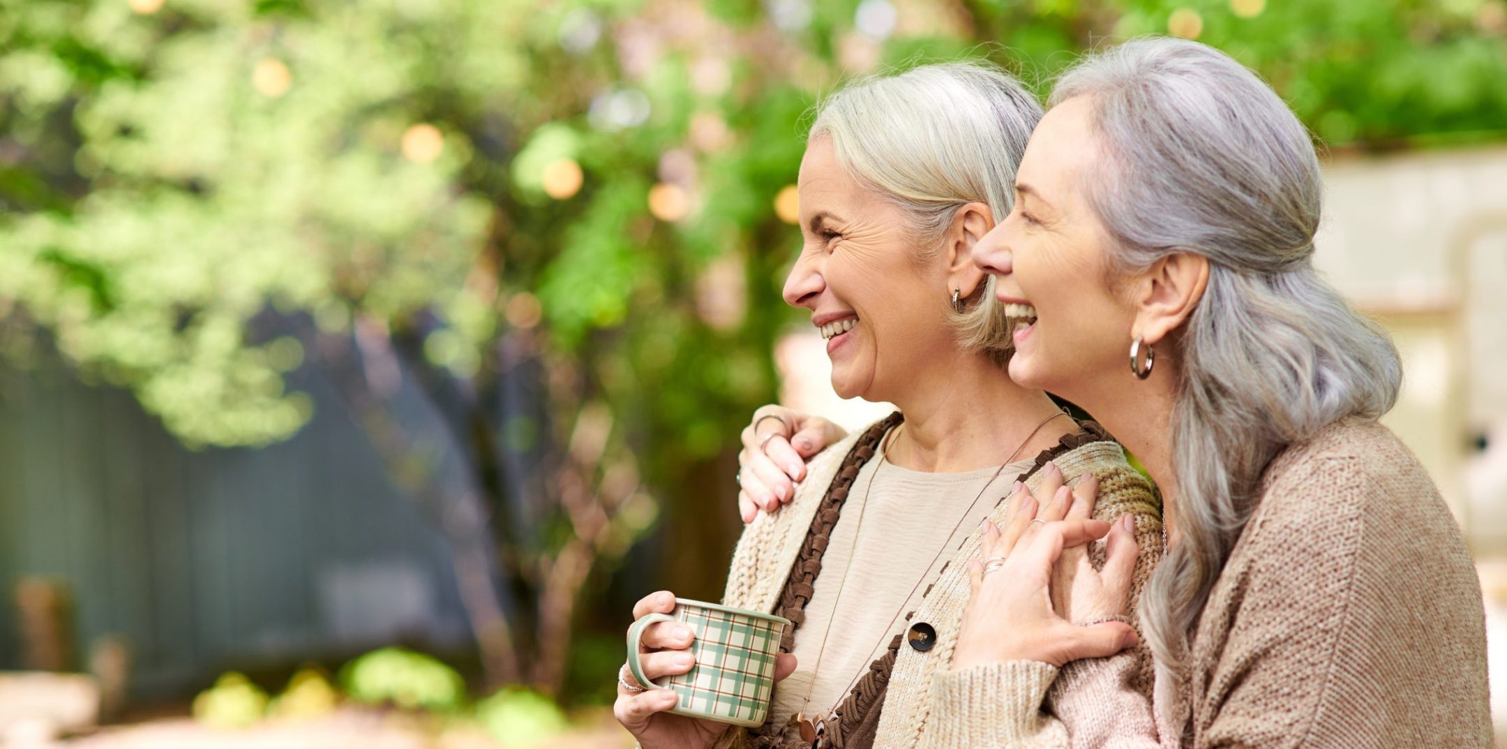 Two older women with gray hair, wearing cozy sweaters, stand in a sunlit garden, smiling and laughing together. One holds a checkered mug, enjoying warm friendship much like the connections built through Eskaton support groups.