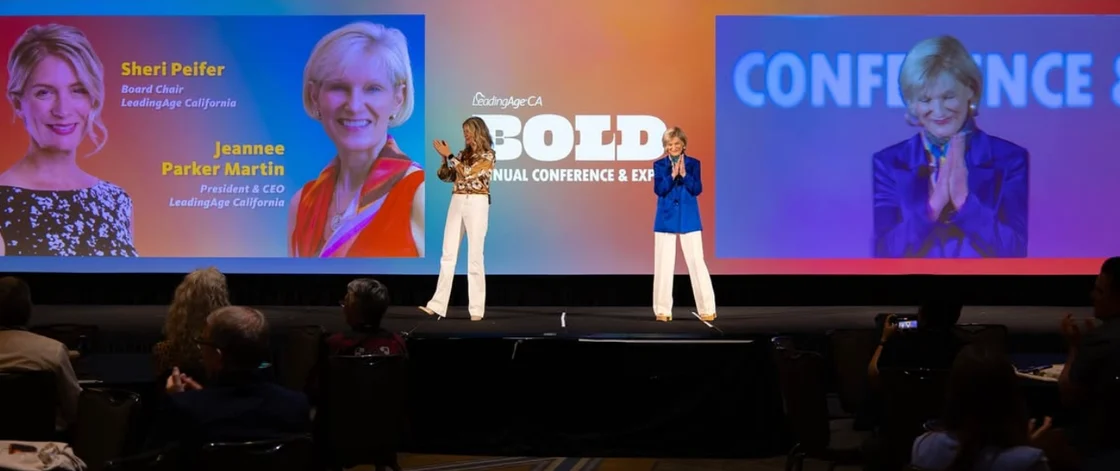 Two women stand onstage speaking at a conference, with a vibrant screen behind them displaying their names and photos: Sheri Peifer and Jeannee Parker Martin. This Eskaton Leadership Through LeadingAge event draws an attentive audience in the foreground.