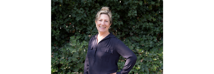 A woman with blonde hair tied up, wearing a long-sleeve black shirt, stands outdoors in front of a leafy green background, smiling at the camera with one hand on her hip—showcasing Eskaton Leadership Through LeadingAge.